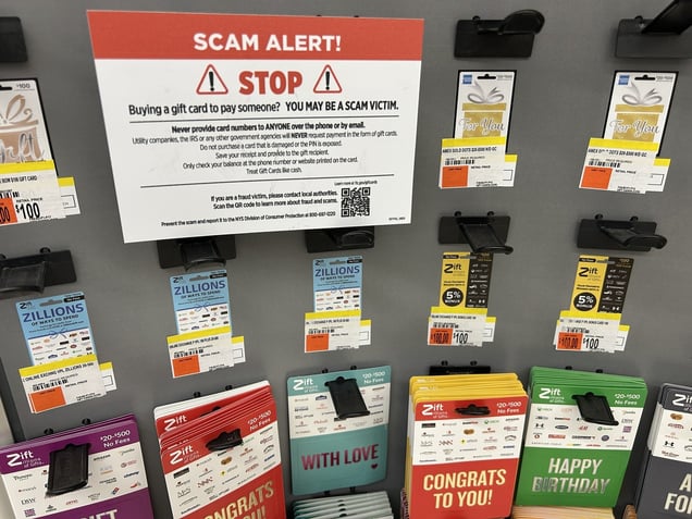 A retail store display of gift cards with a large sign above them warning: "SCAM ALERT! Buying a gift card to pay someone? YOU MAY BE A SCAM VICTIM."
