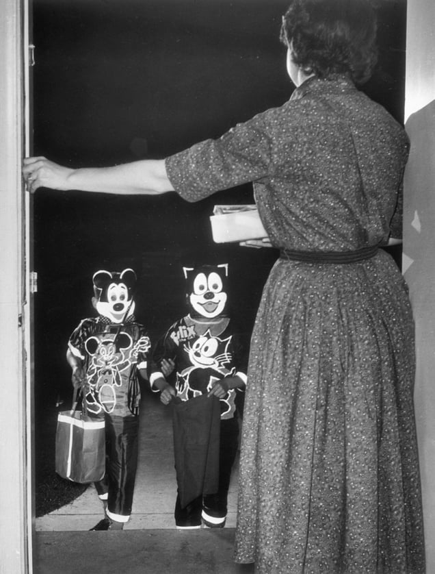 A black-and-white photo from 1960 shows a woman at a doorway giving a treat to two children in Mickey the Mouse and Felix the Cat costumes.