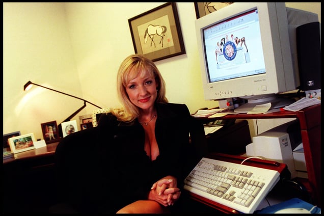 Danni Ashe sits at a desk, wearing a black blazer, with an old-style computer monitor displaying her website visible on the screen.
