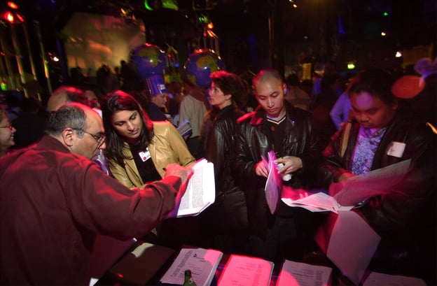 Group of people at a busy indoor event, engaging with documents and each other under dim, colorful lighting; some attendees wear earth-themed hats.