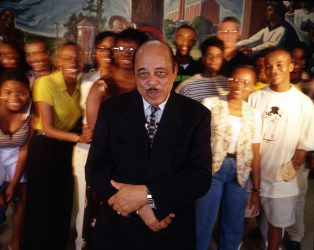 Comer Cottrell, a Black man in a suit, smiles among a diverse, blurred group. 1980s New York City.