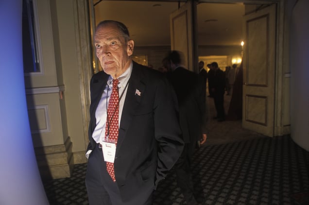 A photo of John Bogle, the founder of Vanguard, wearing a suit and a red patterned tie, standing in a doorway at an event.