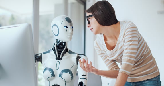A woman in glasses and a striped shirt interacts with a humanoid robot with digital blue eyes in a modern setting.