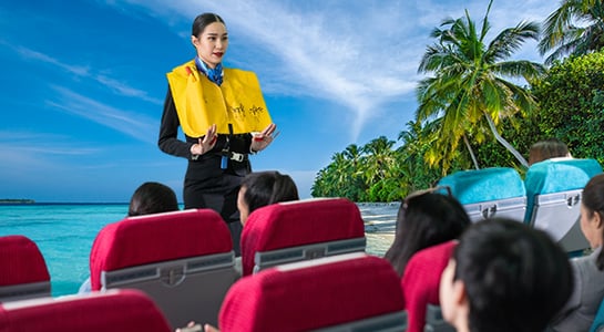 Indoor scene: audience in red chairs, person in yellow life vest addressing them; backdrop of tropical coastal scene, indicating a travel or tourism-related safety briefing.
