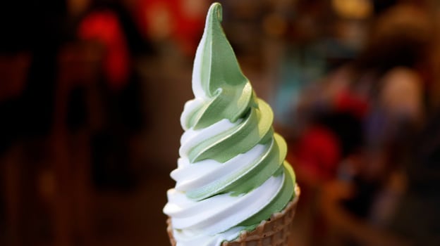 A close-up shot of a swirl of green and white soft-serve ice cream in a waffle cone, with a blurred background.