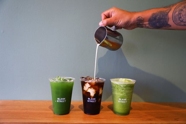 A hand pours milk into an iced drink, with two other Blank Street Coffee beverages on a counter.