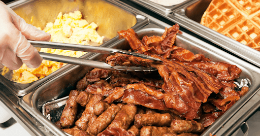 A breakfast buffet pictured with a hand using tongs to pick up bacon.