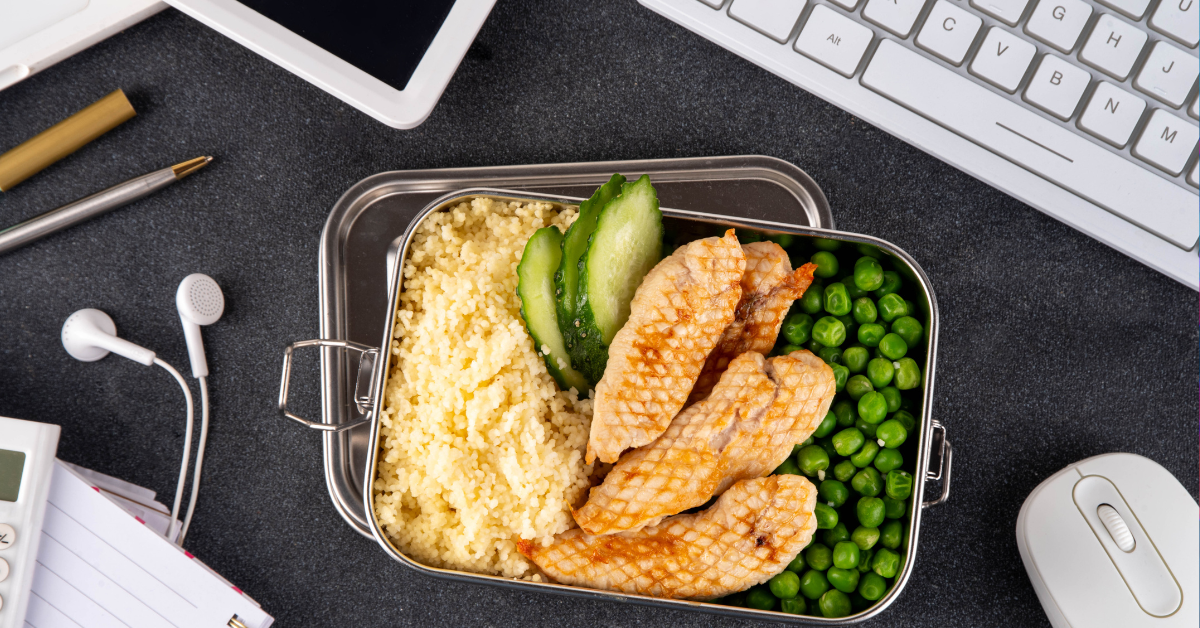  A metal container with peas, chicken, cucumbers, and rice sits on an office desk. Also on the desk are a keyboard, mouse, and earbuds. 