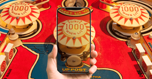 A pinball machine with a hand holding a smartphone in the foreground displaying the same machine on its screen.