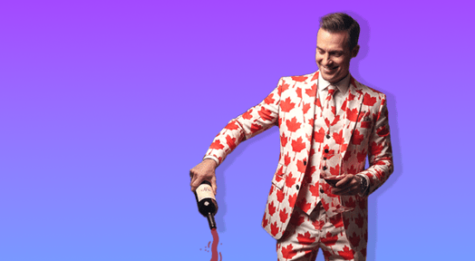 A smiling man in a suit patterned with red Canadian maple leaves pours a stream of wine from a bottle held in his right hand.